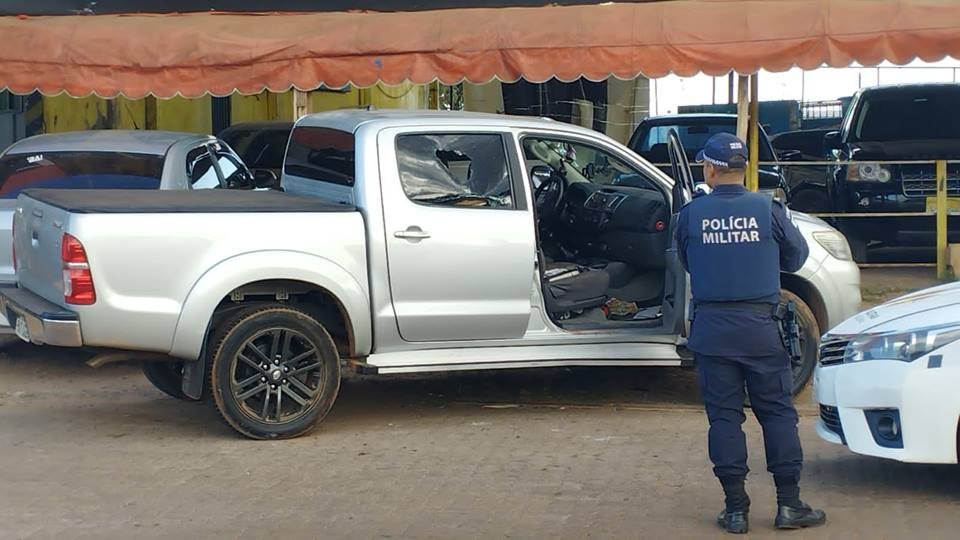 Caminhonete prata com marca de tiro em janela, porta aberta e policial militar