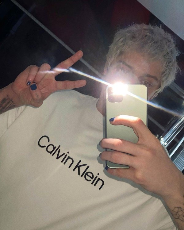 O comediante norte-americano Pete Davidson tirando selfie com o celular no espelho. Ele é um homem branco, com cabelo descolorido, e está posando para foto com uma camiseta branca. Na peça está escrito o nome da marca, Calvin Klein, em letras pretas. 
