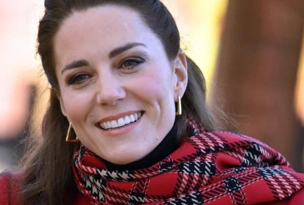 Kate Middleton está usando um cachecol xadrez. Ela sorri e tem cabelos castanhos - Metrópoles