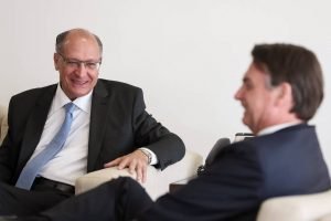Geraldo Alckmin e Bolsonaro conversam e riem. Ambos estão sentados em sofás, lado a lado - Metrópoles