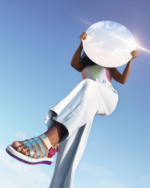 A black woman with long hair is taking pictures outside.  It has a round mirror, which reflects the blue sky and white clouds.  She wears light blue jeans, a colorful body suit and Melissa sandals in white, blue and purple.