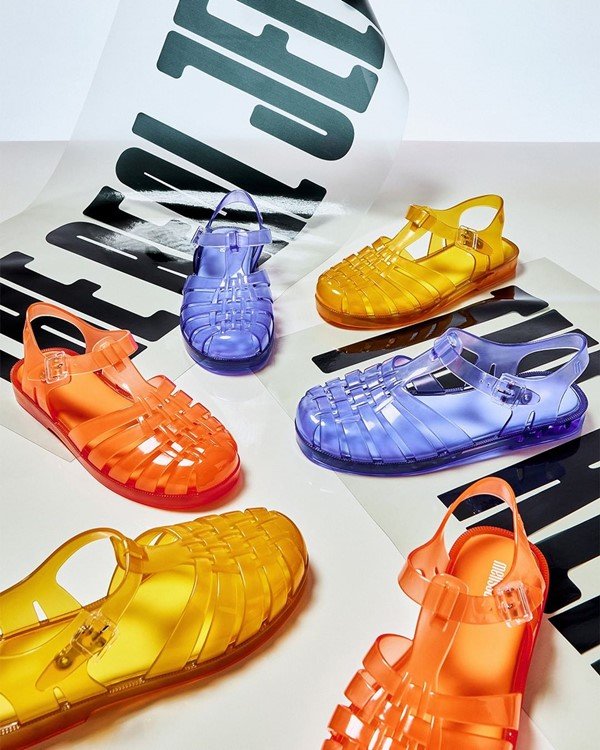 Melissa shoes from the Possession model in yellow, lilac and orange.  Their material is transparent, transparent, in the colors mentioned.  They are all arranged next to each other in a branded exhibition stand.