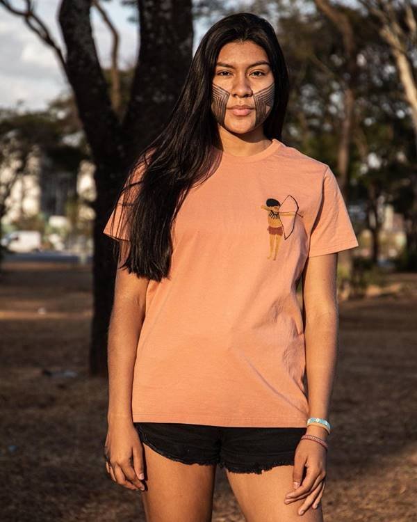 Mulher indígena usa camiseta laranja e shorts preto e está com pintura no rosto