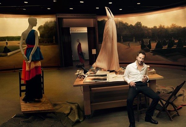 Stylist Tom Ford, a white man with a beard and black hair, sits on a bench and leans against a table where you can see drawings and sketches.  In the background you can see mannequins wearing clothes from the last century. 