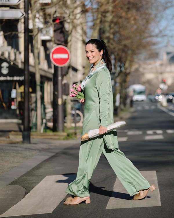 A estilista Chris Gontijo, uma mulher branca com cabelos castanhos lisos e longos, posando para foto com pijama da própria marca em Paris. Na foto, ela usa um conjunto verde água e segura uma baguette, pão típico da França. Ela está atravessando a rua.