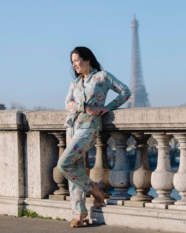 A estilista Chris Gontijo, uma mulher branca com cabelos castanhos lisos e longos, posando para foto com pijama da própria marca em Paris. Na foto, ela usa um conjunto azul com estampa de flores e está escorada em em uma ponte. Ao fundo é possível ver a Torre Eiffel, cartão postal da cidade.
