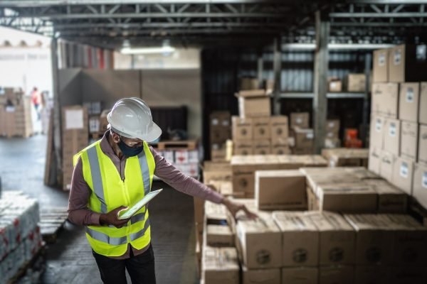homem negro com colete e capacete de trabalho analisa caixas em umas indústria