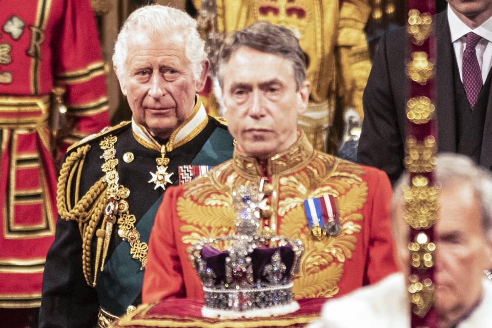Foto colorida. Príncipe Charles ao lado de uma coroa