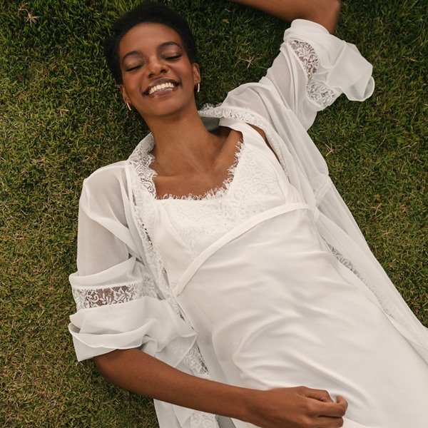 Modelo negra, com cabelos crespo curto, posando para foto deitada na grama verde. Ela usa uma camisola branca com robe, também branco, com detalhes de renda. As peças são da marca Chris Gontijo.