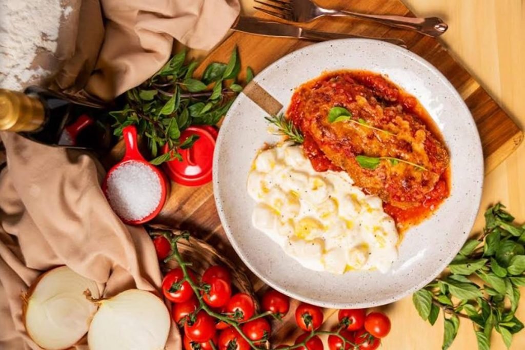 foto de prato com parmegiana e nhoque ao molho branco e ao lado folhinhas verdes e tomatinhos