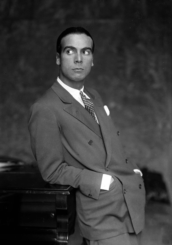 Homem branco, com cabelos escuros penteados para trás, posando para foto encostado em um piano em 1927. Seu nome é Cristóbal Balenciada e ele usa um terno, camisa branca e uma gravata. Ele é o estilista espanhol criador da marca Balenciaga.