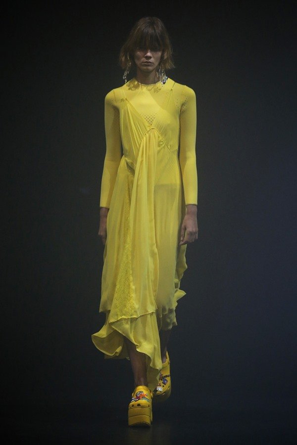 Mujer caucásica, de pelo rubio corto y flequillo, camina por la pasarela Balenciaga.  Lleva un vestido amarillo largo y vaporoso con mangas y zapatos de tacón de piel de cocodrilo del mismo color.