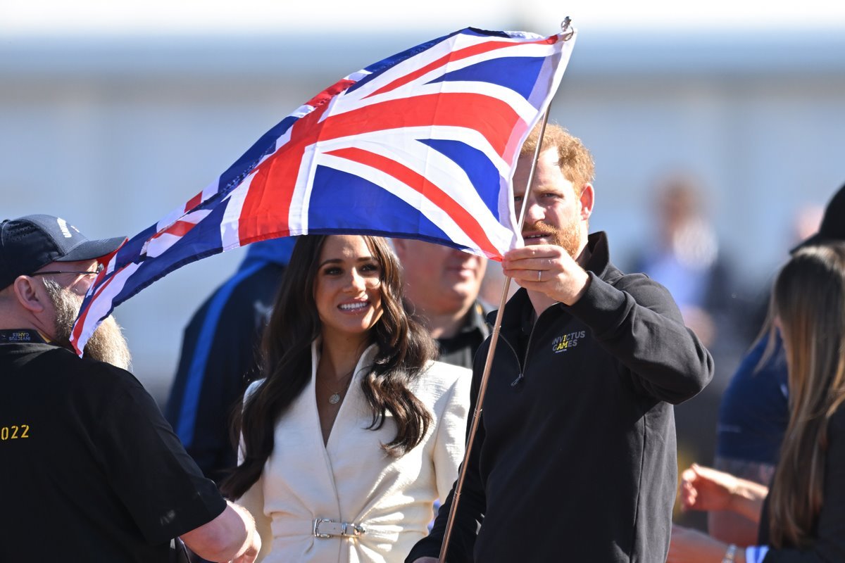 Meghan Markle e princípe Harry em um evento. Ele veste um casaco preto e segura uma bandeira do Reino Unido. Já a atriz veste um blazer branco e está com os cabelos soltos e ondulados.