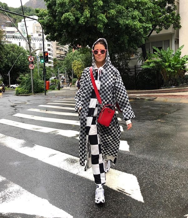 Mulher branca, com cabelos escuros amarrados, atravessando a faixa de pedestre em um dia de chuva. Ela veste casaco e calça com a mesma estampa, um quadriculado branco e preto. Por cima, usa uma capa de chuva na mesma padronagem e uma bolsa transversal vermelha. Seu nome é Lulu Novis e ela é uma influenciadora digital e stylist.