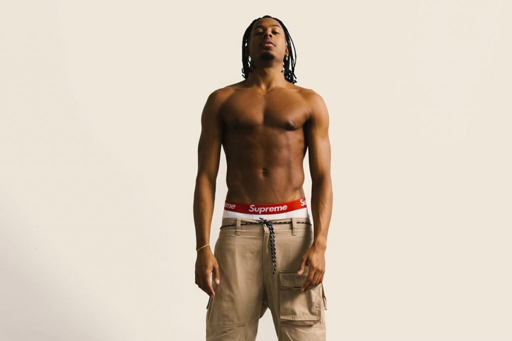 O ex-BBB Paulo André, um homem alto, negro e com cabelos trançados, posando para foto em fundo branco. Ele está sem camisa e usa uma calça aqui com uma cueca branca aparecente. A cueca possui uma faixa vermelha com a logo da empresa, a marca Supreme.