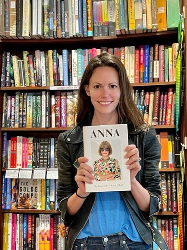 A jornalista Amy Odell em uma livraria, segurando o livro de sua autoria, uma biografia da chefe da Vogue, Anna Wintour. Amy é uma mulher jovem e branca, com cabelos médios castanhos. Ela usa uma blusa azul e jaqueta de couro preta. Ao fundo é possível ver prateleiras com outros livros.