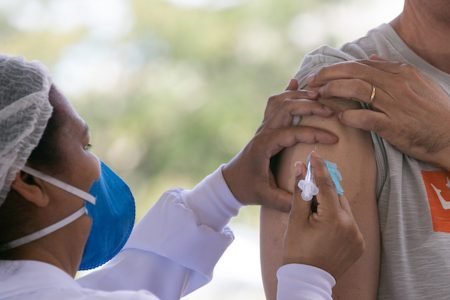 Enfermeira de perfil, com máscara azul PFF e touca na cabeça aplicando vacina em braço de pessoa com camisa cinza