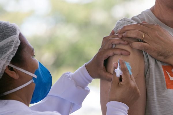 Enfermeira de perfil, com máscara azul PFF e touca na cabeça aplicando vacina em braço de pessoa com camisa cinza