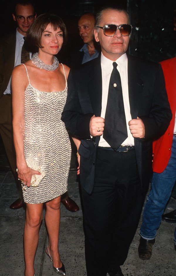 A editora da revista Vogue dos Estados Unidos, Anna Wintour, e o estilista Karl Lagerfeld nas ruas de Nova York em 1990. Ela, uma mulher branca de meia idade, com cabelos curtos, usa um vestido brilhoso prateado e colar na mesma cor. Ele, um homem mais velho, branco e de cabelos grisalhos, usa calça e blazer pretos e uma camisa branca.