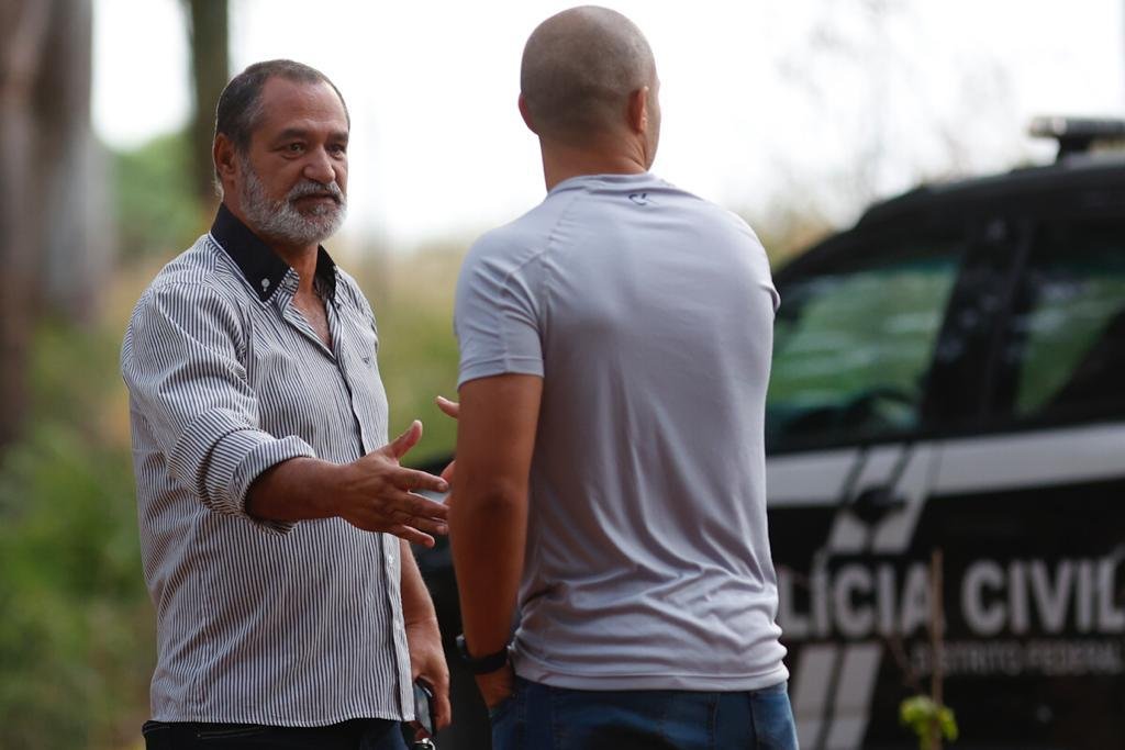 Homens de camiseta cinza apertando a mão um do outro. Ao fundo, viatura preta da Polícia Civil