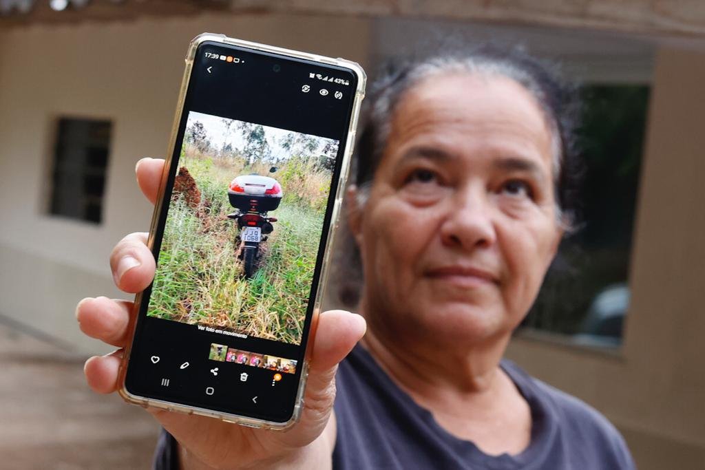 Mulher com cabelo preso olhando para o alto e mostrando foto de moto em matagal em celular