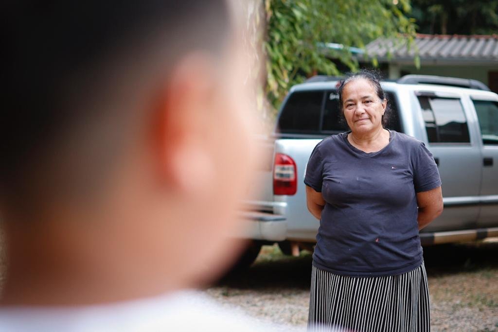 Rosto de criança de costas e desfocado e mulher ao fundo com camisa escura e as mãos para trás