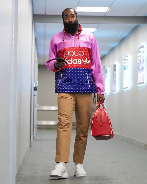Jogador com barba volumosa usa casaco da colaboração adidas x gucci nos tons azul, vermelho e rosa, calça na cor caramelo e carrega bolsa vermelha na mão