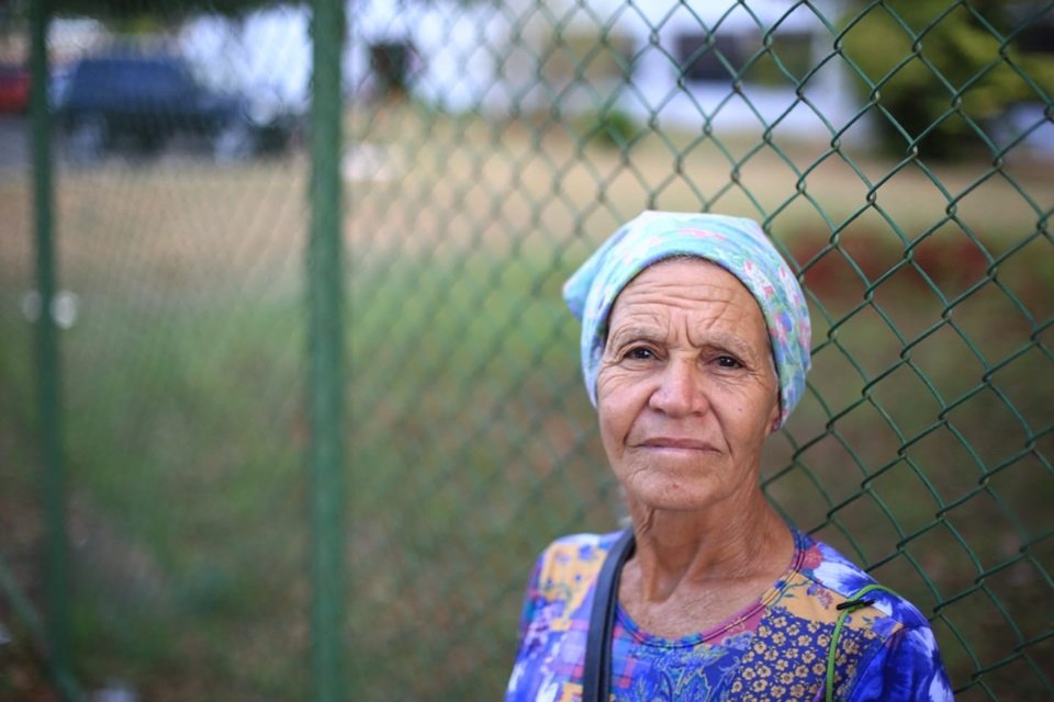 Mulher idosa com pano na cabeça e blusa azul encostada em grade verde, em frente a gramado