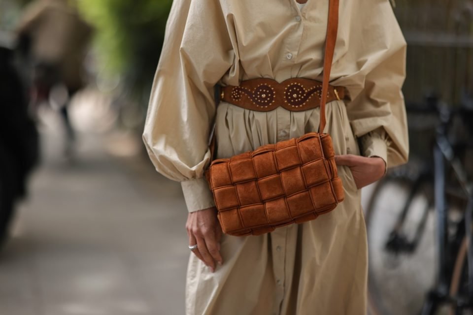 Mulher branca andando na rua e usando um vestido midi bege da SoSue, um cinto de couro marrom Yves Saint Laurent e uma bolsa de couro de camurça marrom Bottega Veneta Cassette.