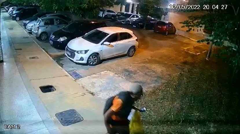 Homem com gorro carrega tampa de bueiro. No fundo um carro branco. Foto colorida