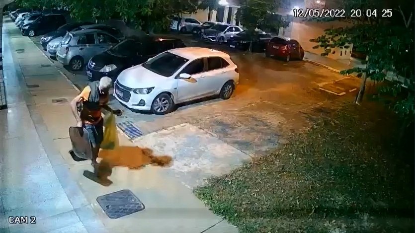 Homem tira tampa de bueiro da rua. Usa gorro e camisa preta. No fundo um carro branco. Foto colorida