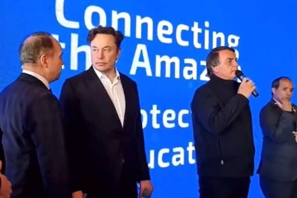 O bilionário Elon Musk, empresário sul-africano ao lado do presidente Jair Bolsonaro, que fala ao microfone em evento do encontro dos dois. Ao fundo, um painel azul e branco - Metrópoles