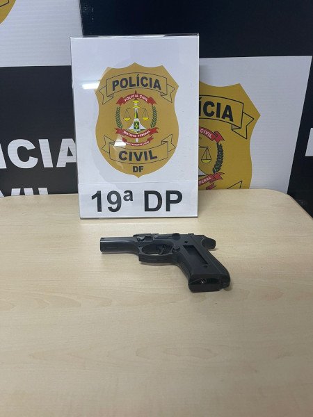 simulacro de arma em cima de mesa de madeira com logo da polícia civil atrás