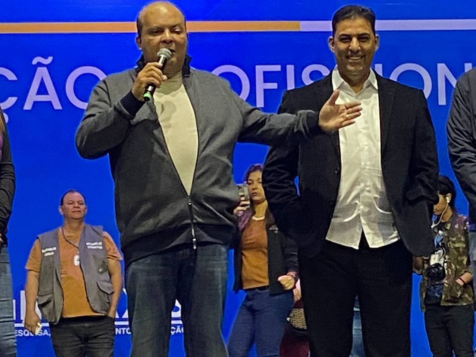 dois homens na frente de um fundo azul. um deles com os braços abertos. em uma das mãos há um microfone
