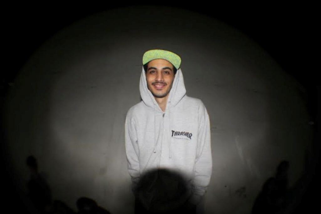 Homem branco com boné verde veste casaco com capuz cinza. Fundo cinza