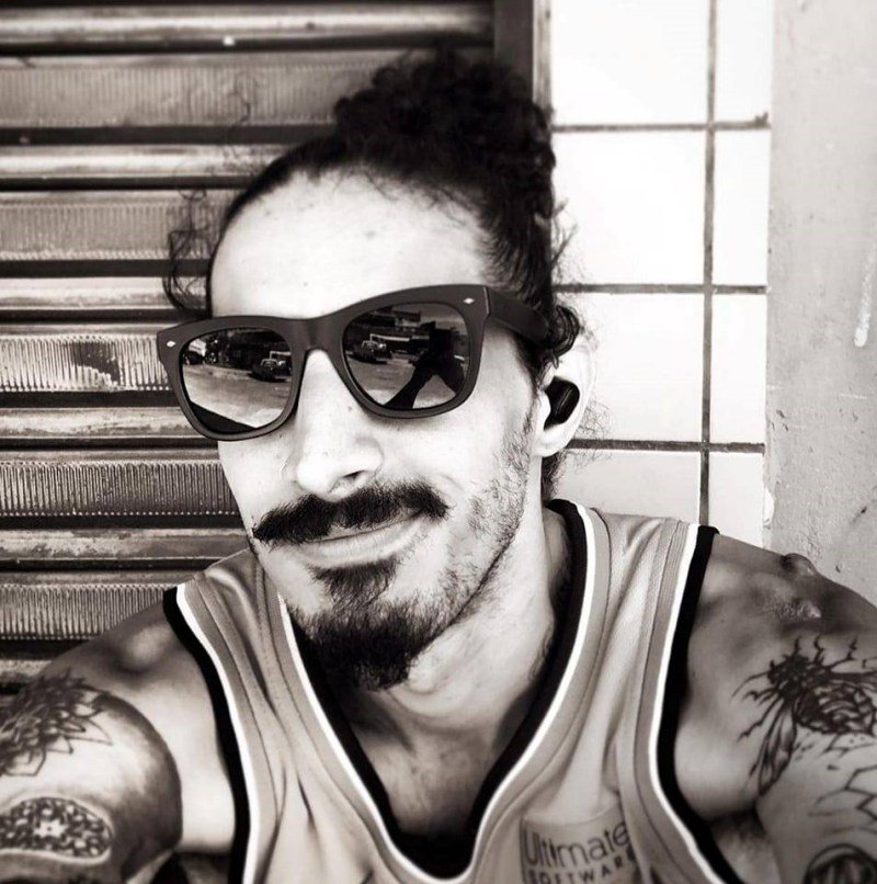 Foto em preto e branco. Homem de bigode e cavanhaque veste camisa regata. Também usa óculos escuros e possui cabelo preso