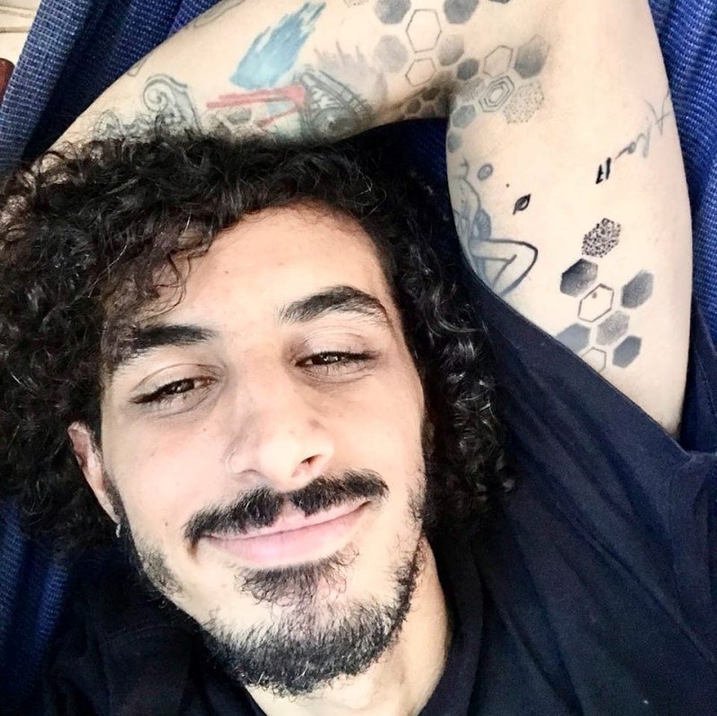 Homem deitado com braços tatuados acima da cabeça. Sorri levemente para a foto. Veste camisa azul escura