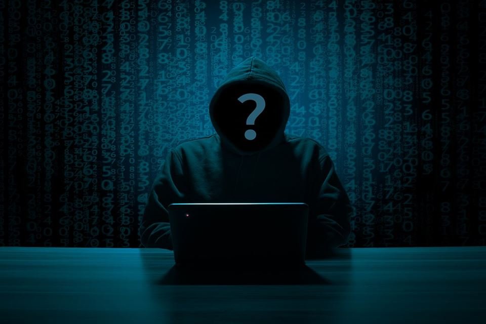 Hacker diante de computador com capuz e interrogação em seu rosto, sob fundo com números em azul - Metrópoles