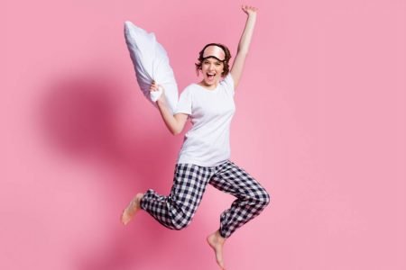 Mulher de pijama com travesseiro na mão pulando em frente a fundo rosa