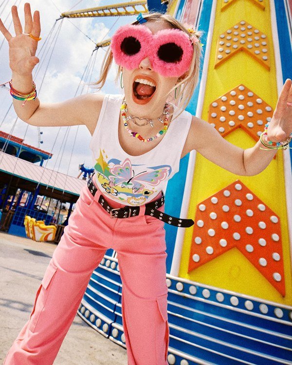 Mulher branca com cabelos louros posando para foto em um parque de diversões. Ela veste roupas da marca chinesa Shein: uma calça rosa, cinto preto com taxas, uma camiseta branca e óculos rosa de pelúcia.