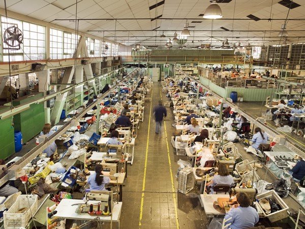 Foto de uma fábrica de roupas. Pessoas estão sentadas em mesas trabalhando nas máquinas de costura 