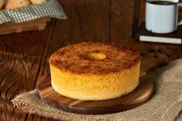 foto de bolo de milho cremoso sobre tabua de madeira com fundo de madeira, paes de queijo e caneca com café