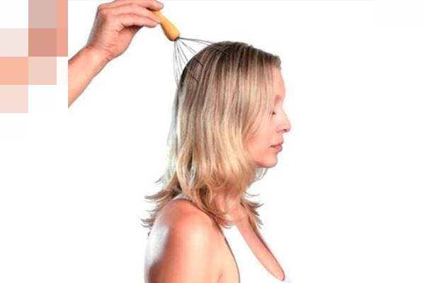 Fotografia colorida de mão segurando massageador sobre cabeça de uma mulher loira que está de olhos fechados