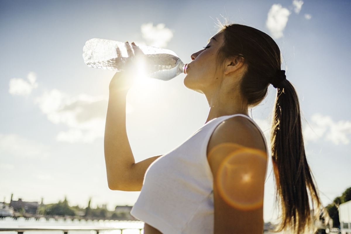 Mulher bebendo água após praticar exercícios físicos, contra a luz do sol. Ela é branca e tem rabo de cavalo - Metrópoles