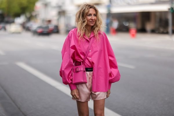 Mulher loira, no street style, usa look todo rosa, com camisa social oversized, shorts de cetim e bolsa pequena transpassada