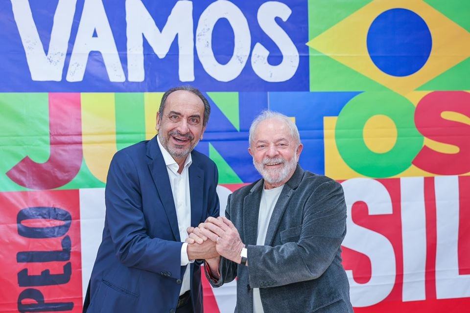 O prefeito de Belo Horizonte e pré-candidato ao governo mineiro, Alexandre Kalil, dá as mãos ao ex-presidente Lula diante de banner de sua campanha. Ambos sorriem e olham para a câmera - Metrópoles