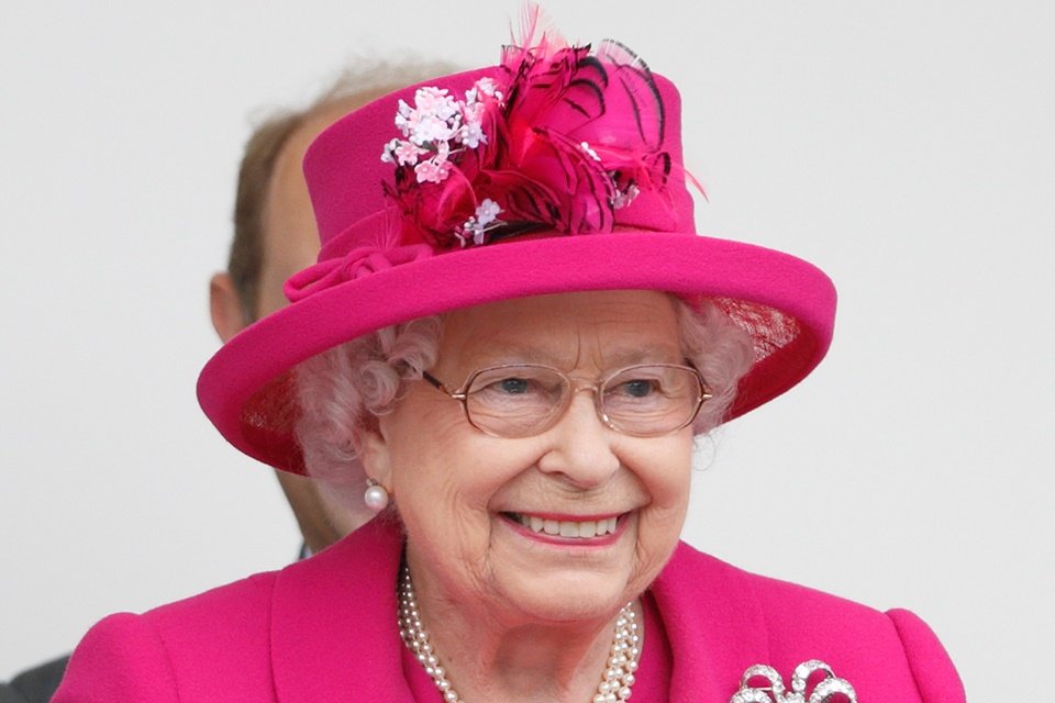 Rainha Elizabeth II sorrindo em evento. Ela usa chapéu com adornos e blazer, ambos rosa, além de colar de pérolas e óculos - Metrópoles