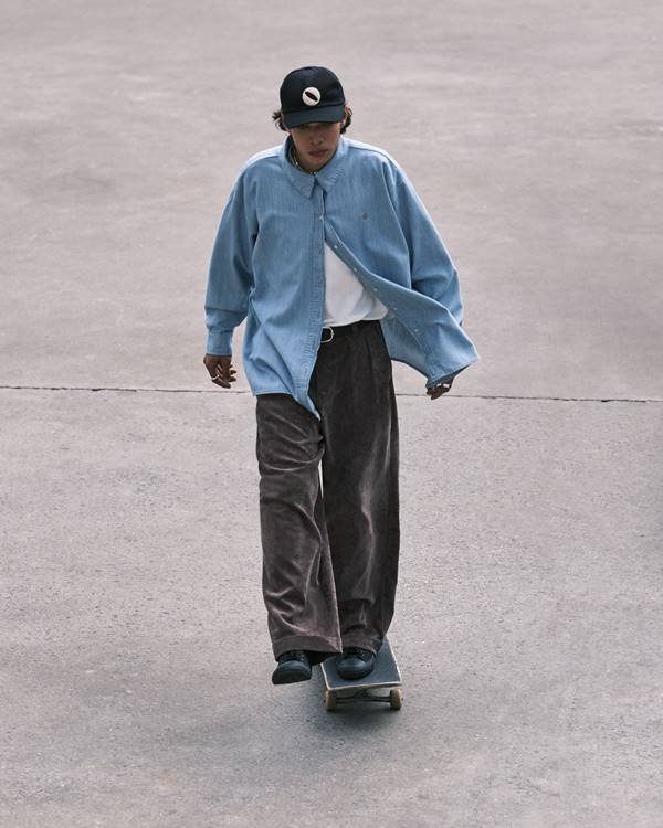 modelo em cima de um skate usa calça cinza, camiseta branca, camiseta jeans e boné