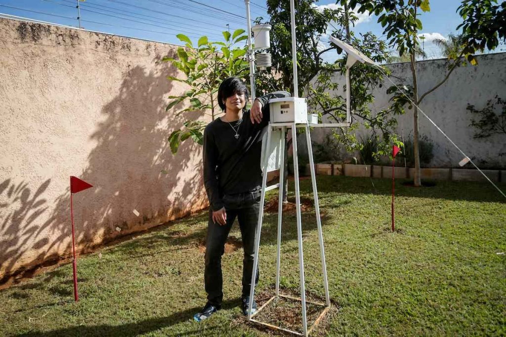 Homem jovem de cabelos pretos apoia-se em estação meteorológica no quintal