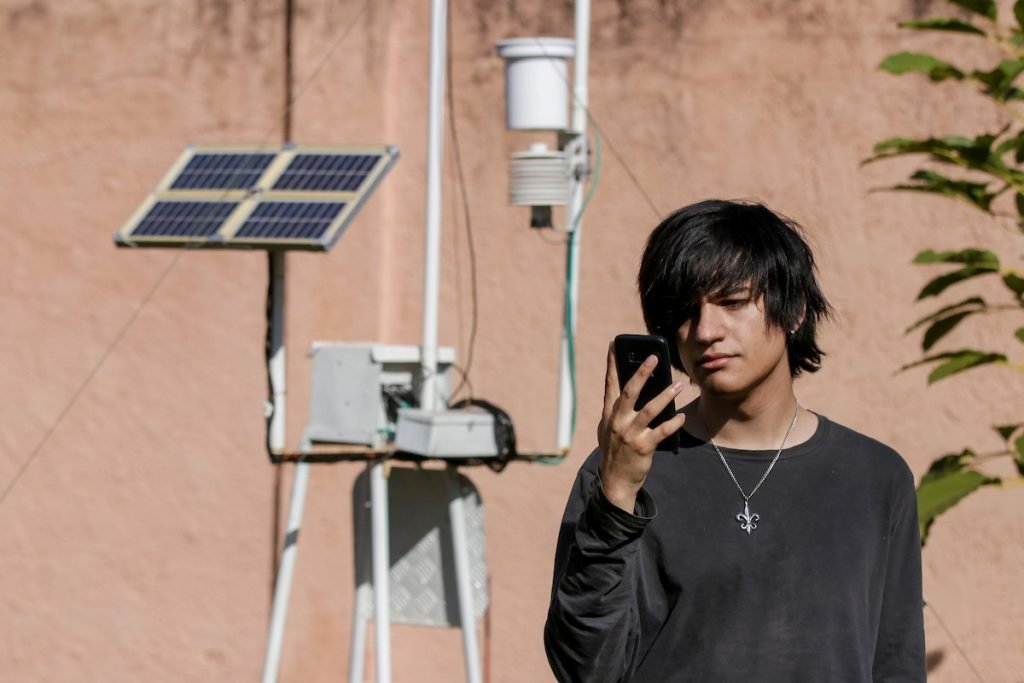 Homem jovem de cabelos pretos seguros celular ao lado de estação meteorológica
